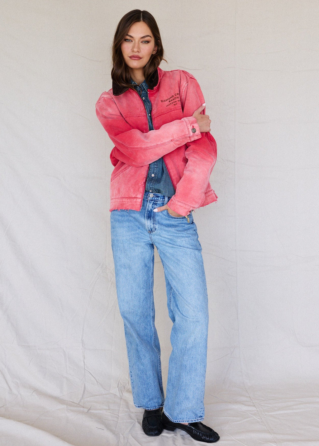 Faded Red | Woman wearing a pink jacket and blue jeans against a plain background