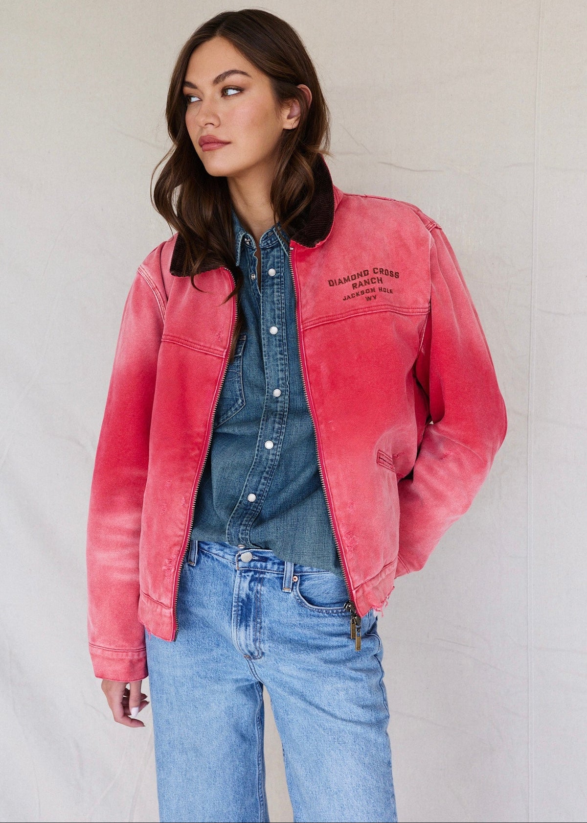 Faded Red | Woman wearing a pink jacket over a denim shirt and jeans against a plain background