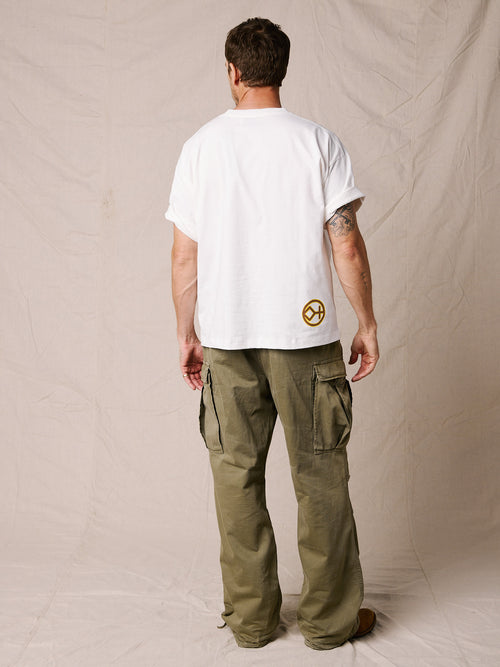 White | A man modeling a white short-sleeve t-shirt with a brown 'WYOMING' graphic and cowboy silhouette, paired with olive green cargo pants and brown boots, standing against a beige fabric backdrop.