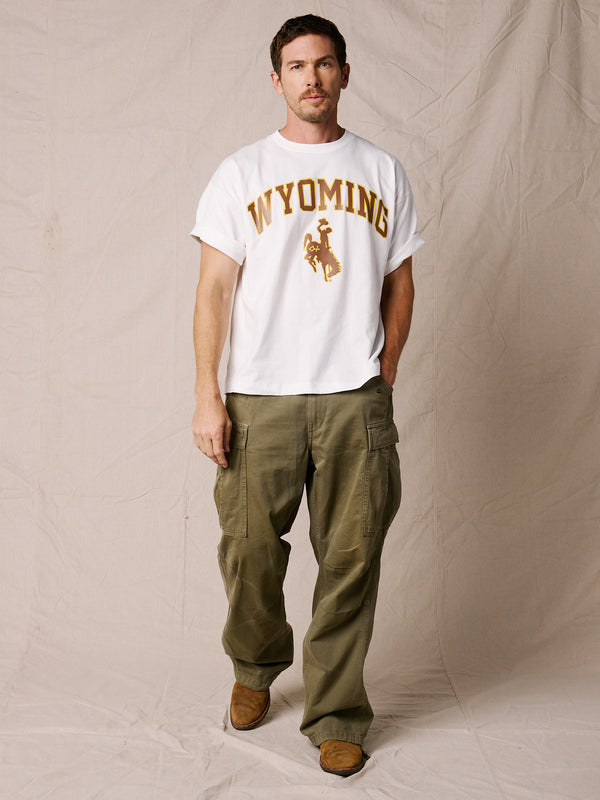 White | A man modeling a white short-sleeve t-shirt with a brown 'WYOMING' graphic and cowboy silhouette, paired with olive green cargo pants and brown boots, standing against a beige fabric backdrop.