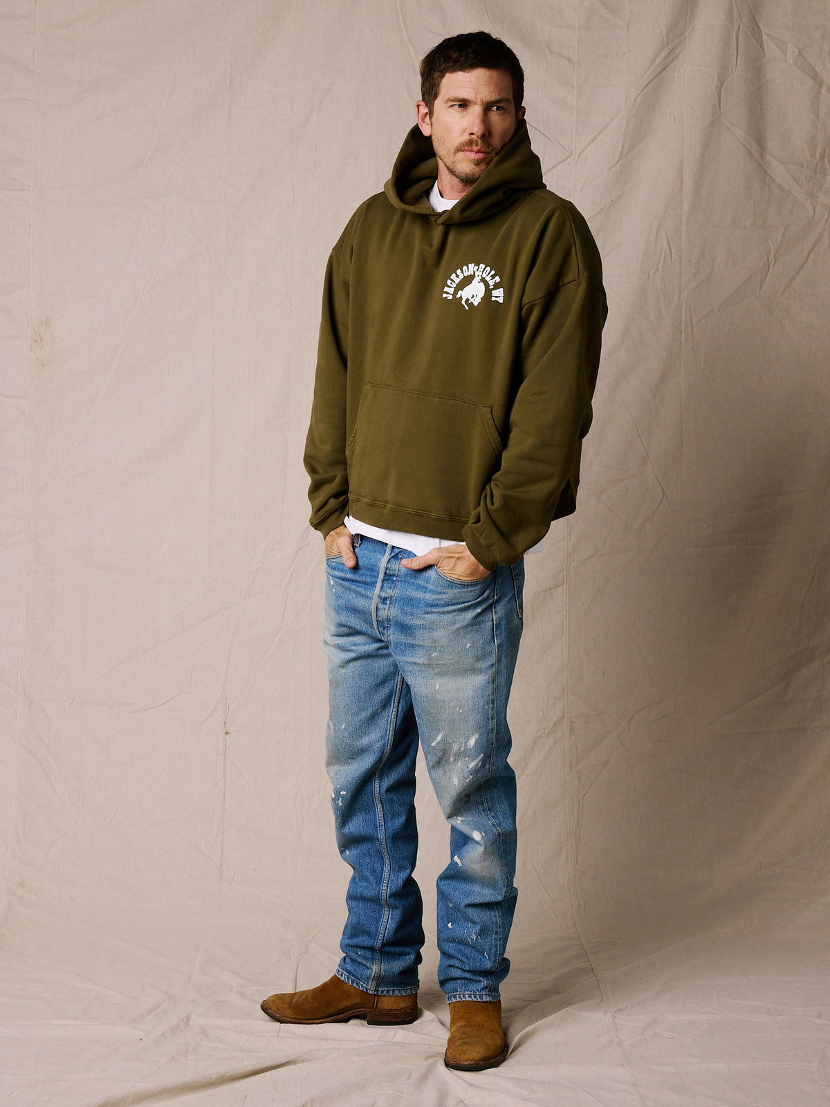 Army Green | A man wearing an olive green pullover hoodie with a white graphic logo on the left chest, paired with blue jeans and brown boots, standing against a beige fabric backdrop.