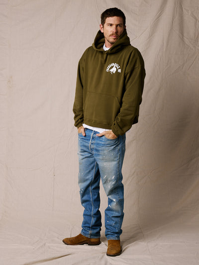 Army Green | A man wearing an olive green pullover hoodie with a white graphic logo on the left chest, paired with blue jeans and brown boots, standing against a beige fabric backdrop.