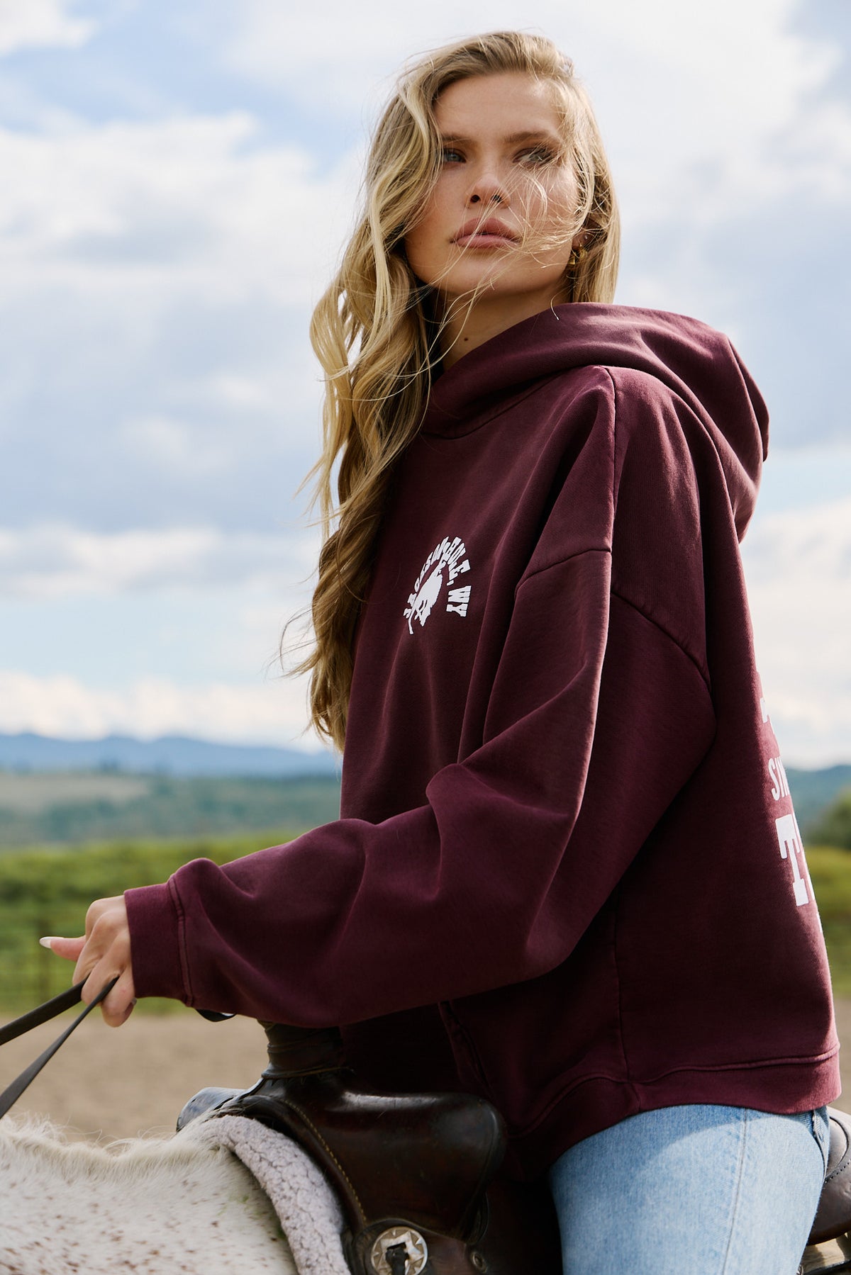 Burgundy | Woman wearing a maroon hoodie with a logo, standing outdoors with a scenic background.