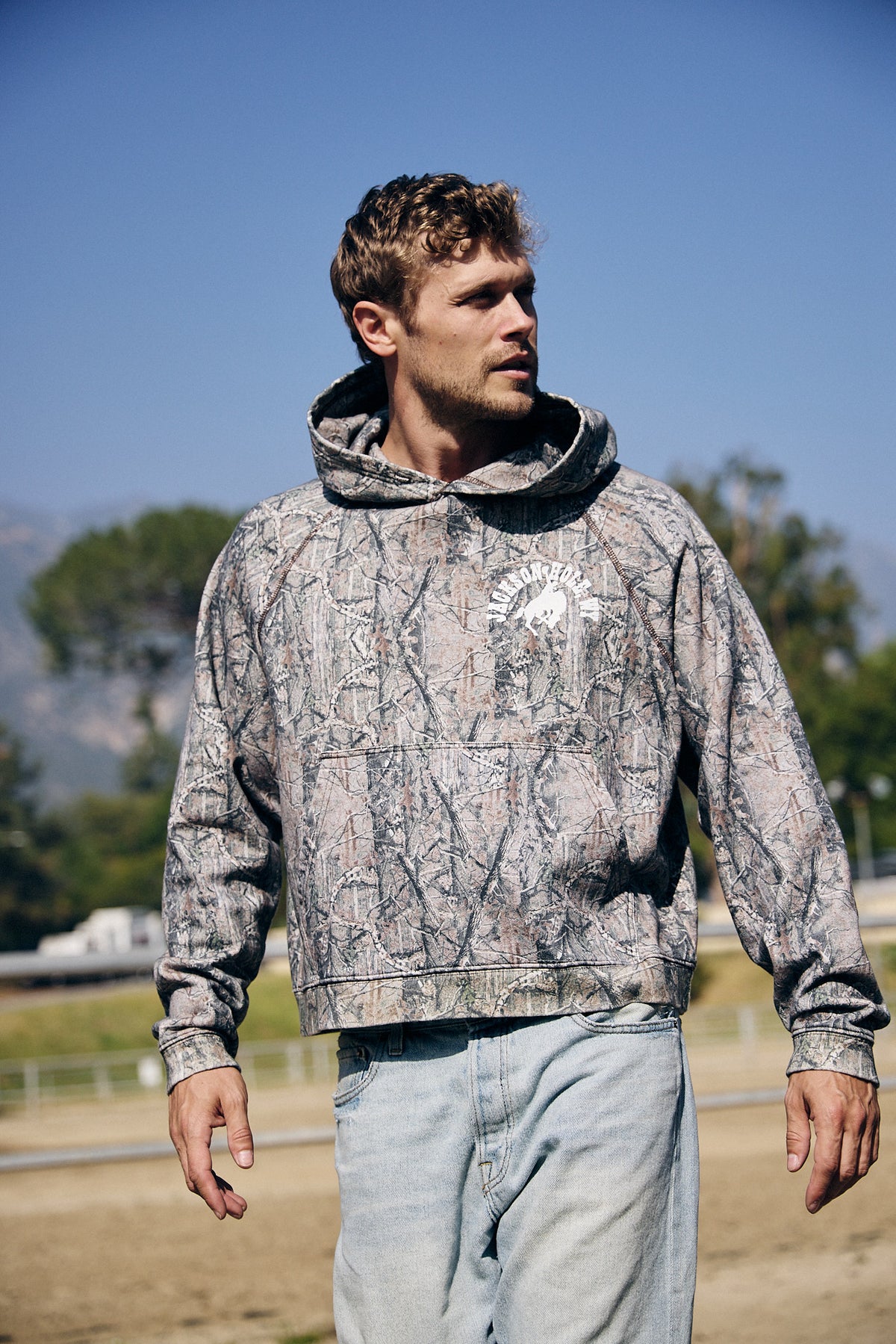 Sage Brush Camo | Man wearing a camouflage hoodie outdoors with trees and clear sky in the background