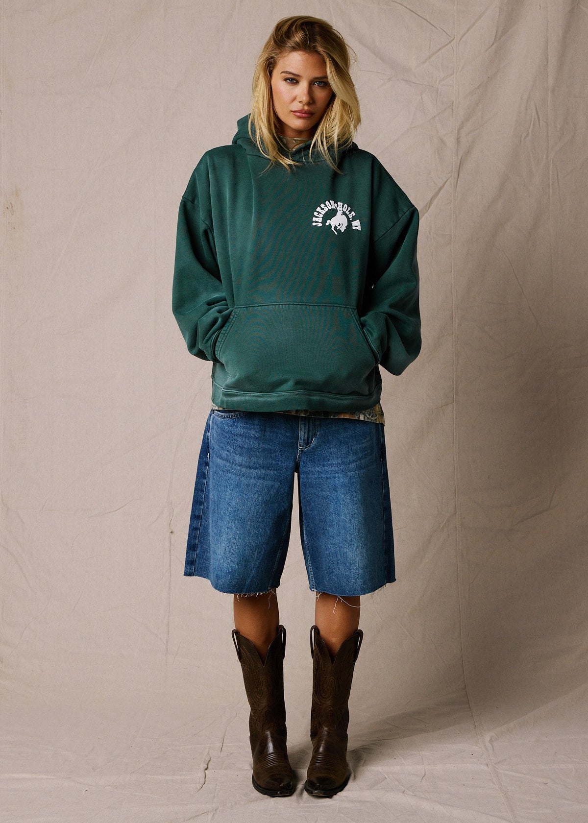 Sun Faded Green | A woman stands against a beige fabric backdrop wearing an oversized dark green hoodie with a white western-style logo on the chest, blue denim shorts, and dark brown cowboy boots. 