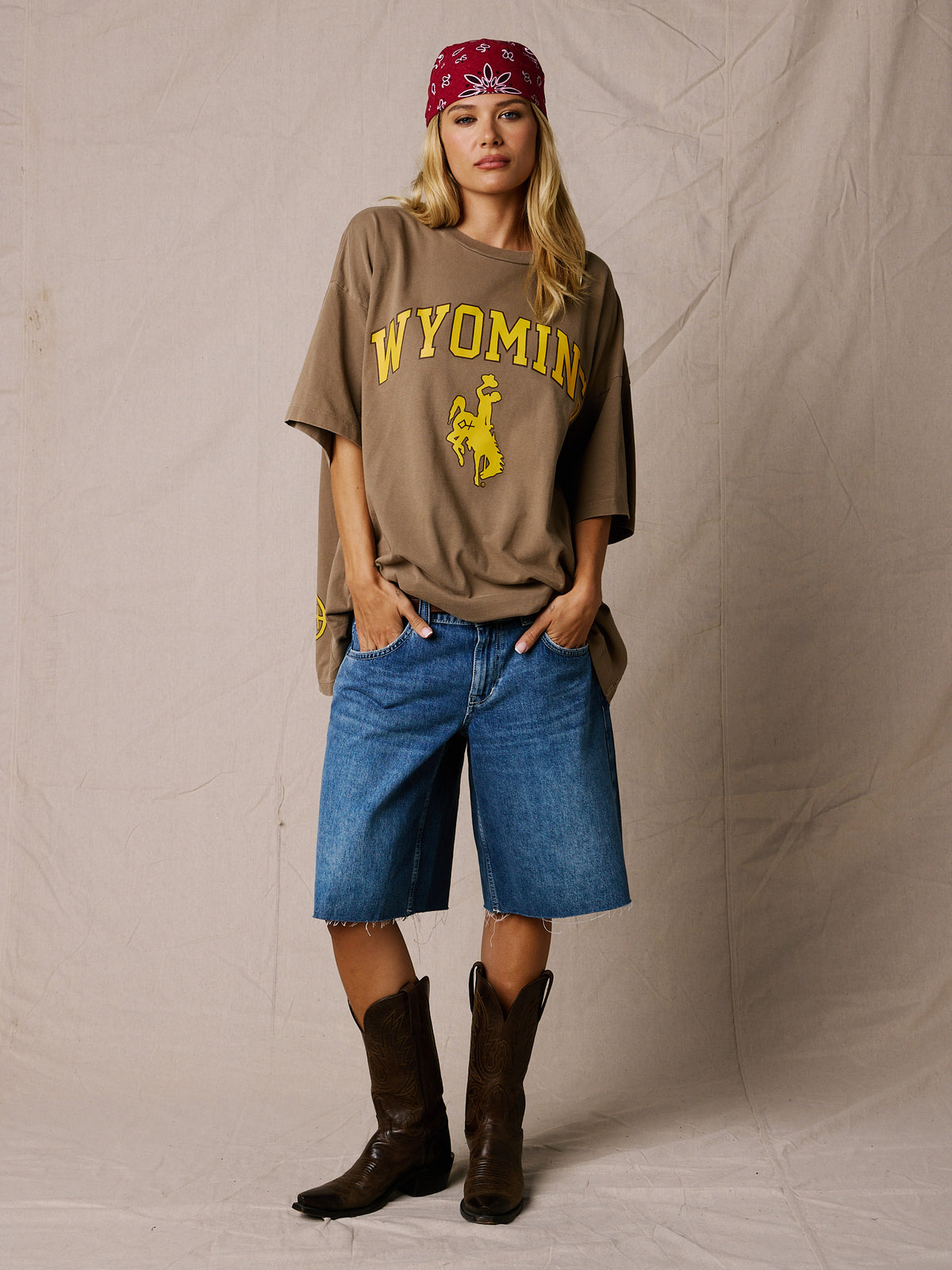Vintage Brown | A woman stands against a wrinkled beige backdrop wearing an oversized brown graphic T-shirt with yellow 'WYOMING' text and a bucking horse logo, blue denim shorts, and dark brown cowboy boots. She also wears a red bandana.