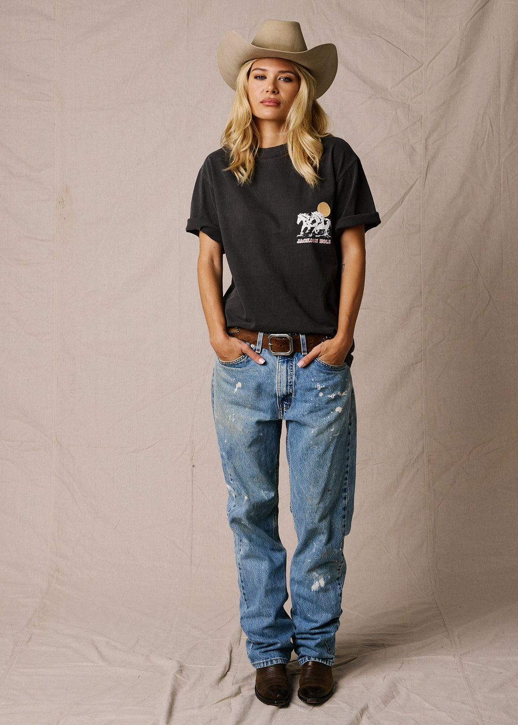 Faded Black | A woman stands against a beige fabric backdrop wearing a black graphic T-shirt, light blue distressed jeans, a tan cowboy hat, and dark brown boots. The T-shirt has a small white and yellow graphic on the left chest.