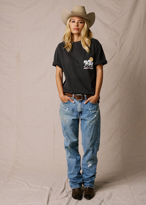 Faded Black | A woman stands against a beige fabric backdrop wearing a black graphic T-shirt, light blue distressed jeans, a tan cowboy hat, and dark brown boots. The T-shirt has a small white and yellow graphic on the left chest.
