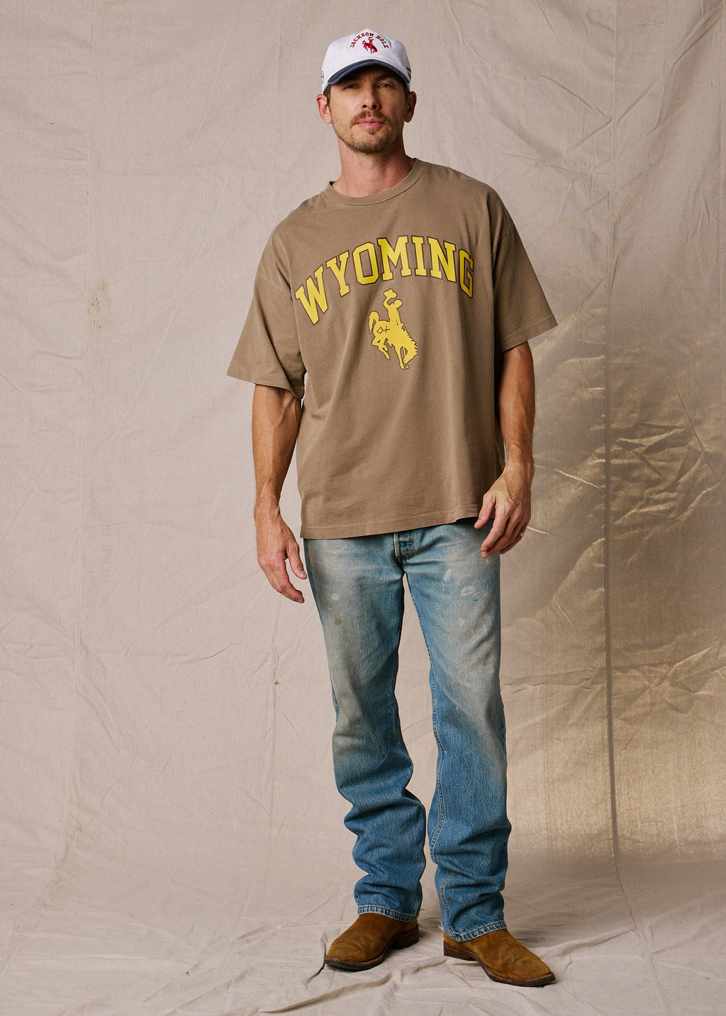 Vintage Brown | A man modeling a brown short-sleeve t-shirt with a yellow 'WYOMING' and bucking horse graphic, paired with blue jeans, brown boots, and a white baseball cap, standing against a beige fabric backdrop.