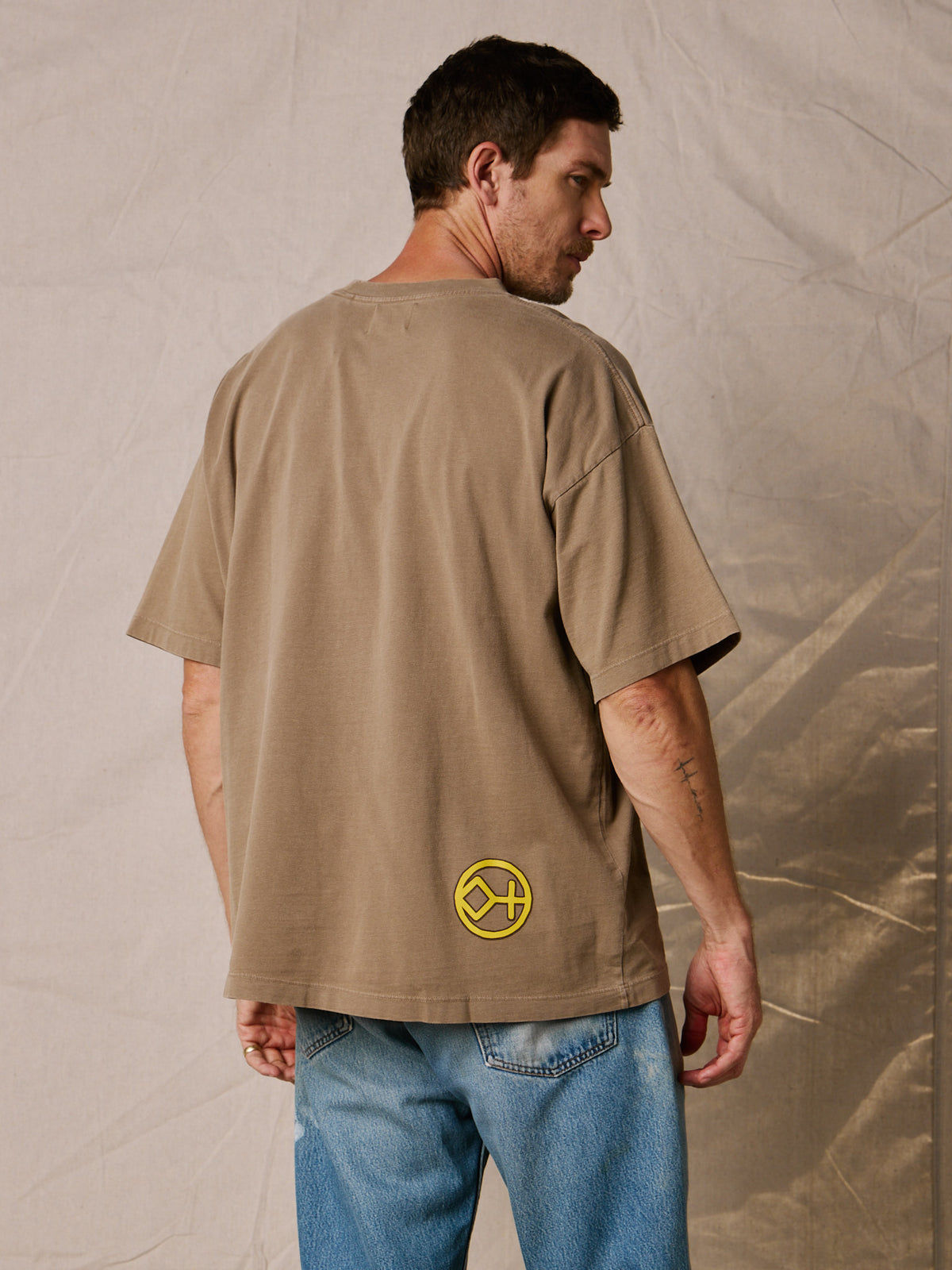 Vintage Brown | A man wearing an oversized tan t-shirt with a yellow circular logo on the lower back, paired with blue jeans, photographed against a neutral textured backdrop.