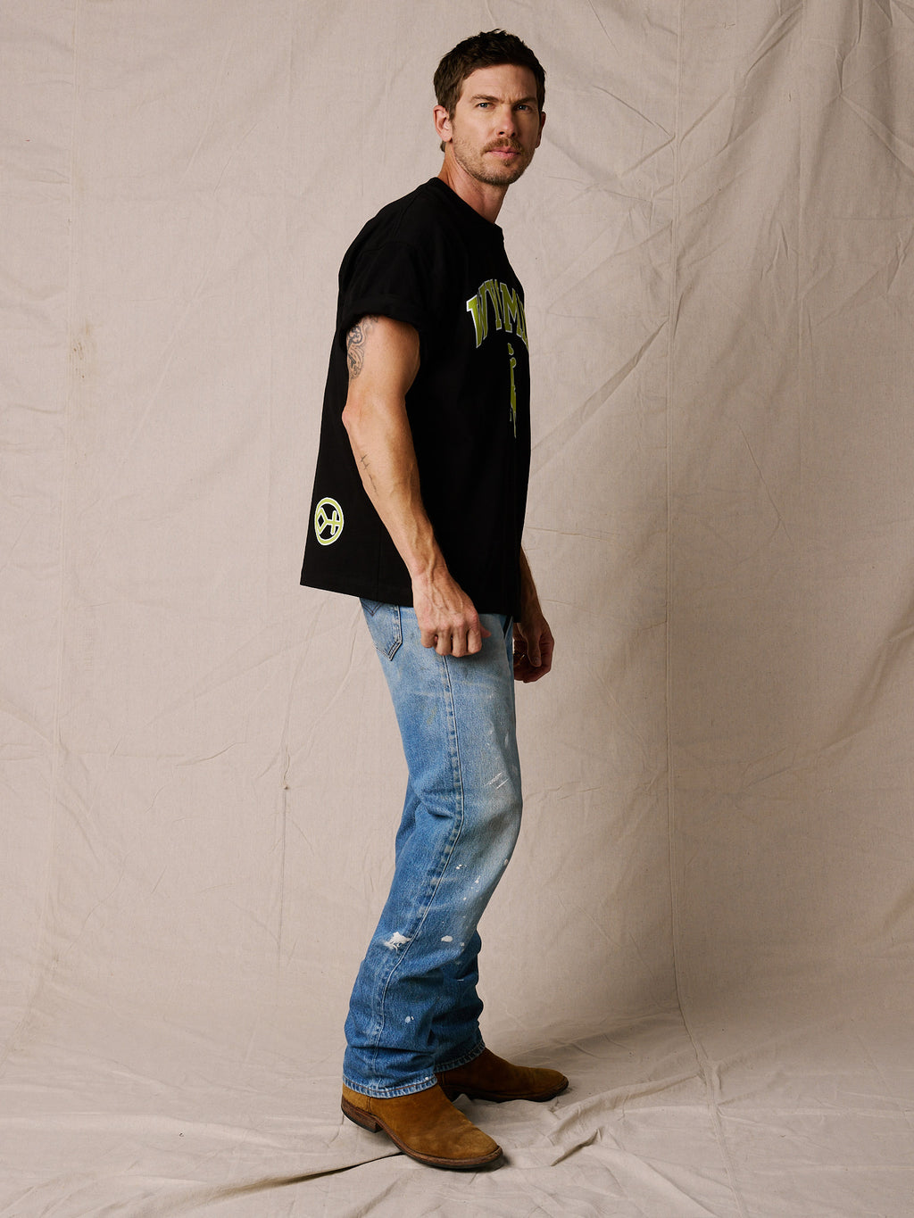 Black | A man modeling a black graphic t-shirt with green and yellow lettering, paired with blue jeans and brown boots, standing against a beige fabric backdrop.
