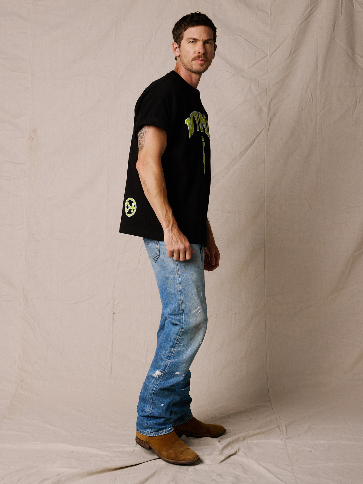 Black | A man modeling a black graphic t-shirt with green and yellow lettering, paired with blue jeans and brown boots, standing against a beige fabric backdrop.