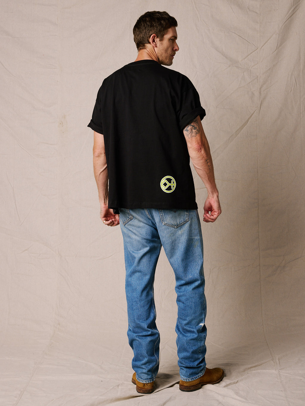 Black| A man viewed from the back wearing a black t-shirt with a small green circular logo on the lower back, paired with blue jeans and brown boots, standing against a beige fabric backdrop.