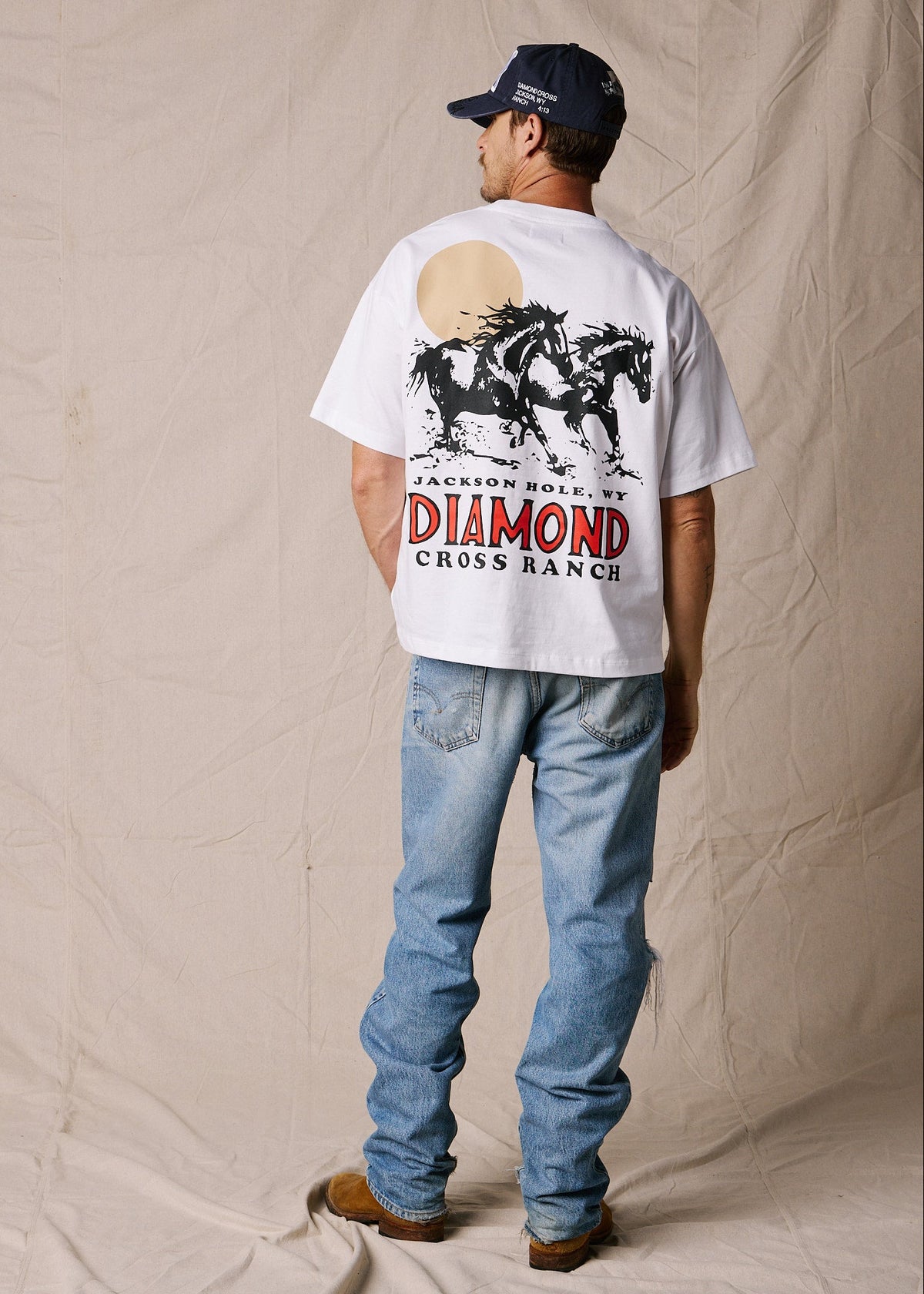Bright White | A man stands with his back to the camera wearing a white graphic t-shirt with a large western-style horse print and 'Jackson Hole WY Diamond Cross Ranch' text, paired with blue jeans and a navy baseball cap, against a beige fabric backdrop.