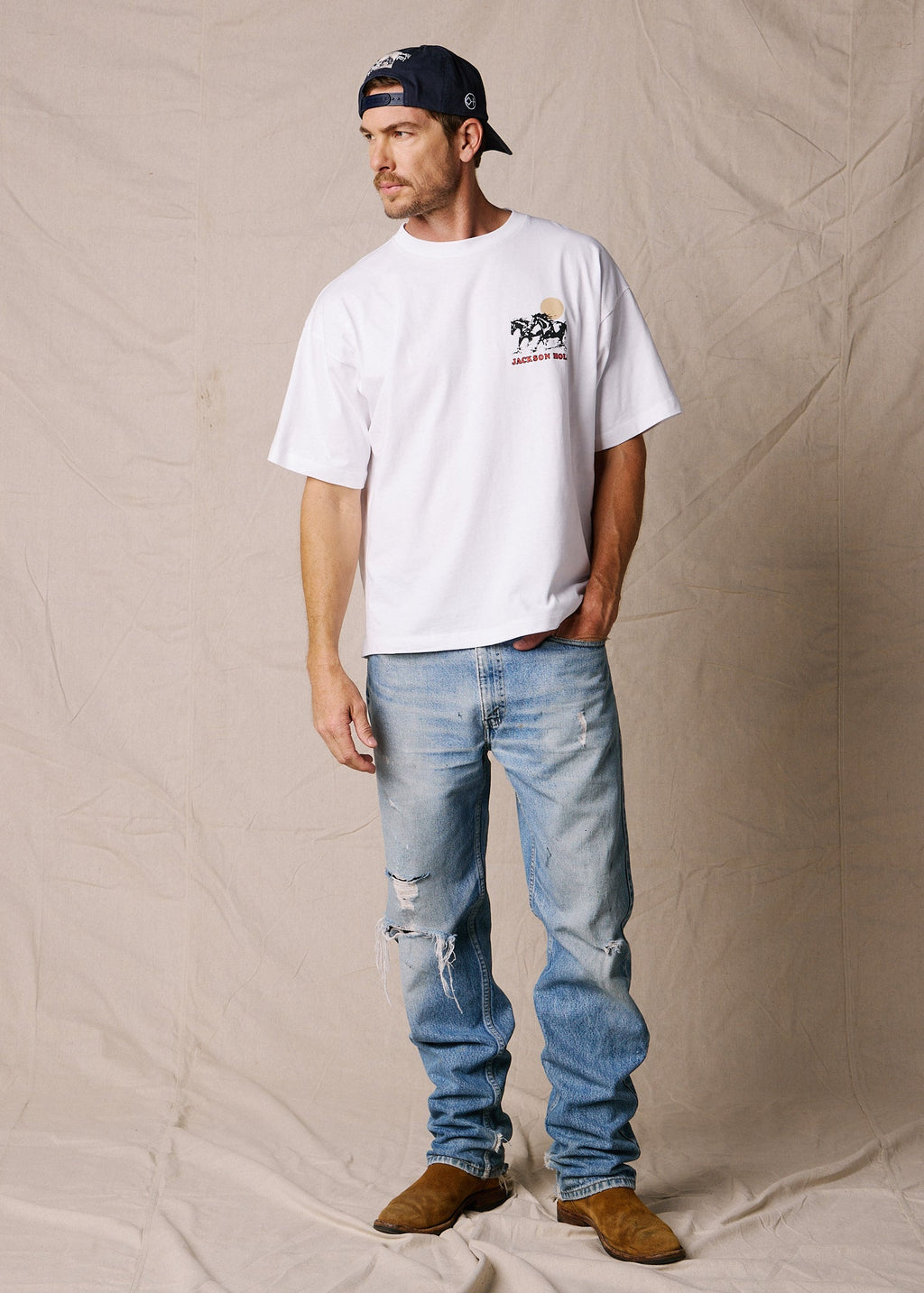 Bright White | A man wearing a white crewneck t-shirt with a small graphic on the left chest, light blue distressed jeans, brown suede boots, and a dark baseball cap, standing against a beige fabric backdrop.