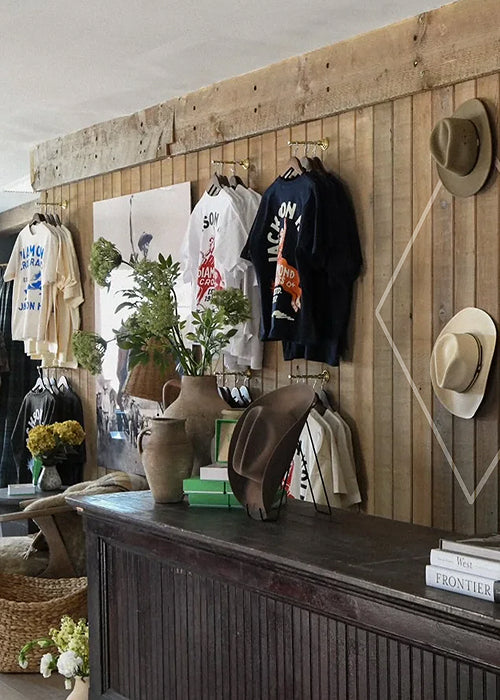 A rustic clothing display in a shop featuring graphic t-shirts on hangers, a dark brown wide-brim hat on a wooden counter, and two straw fedora hats hanging on the wood-paneled wall.